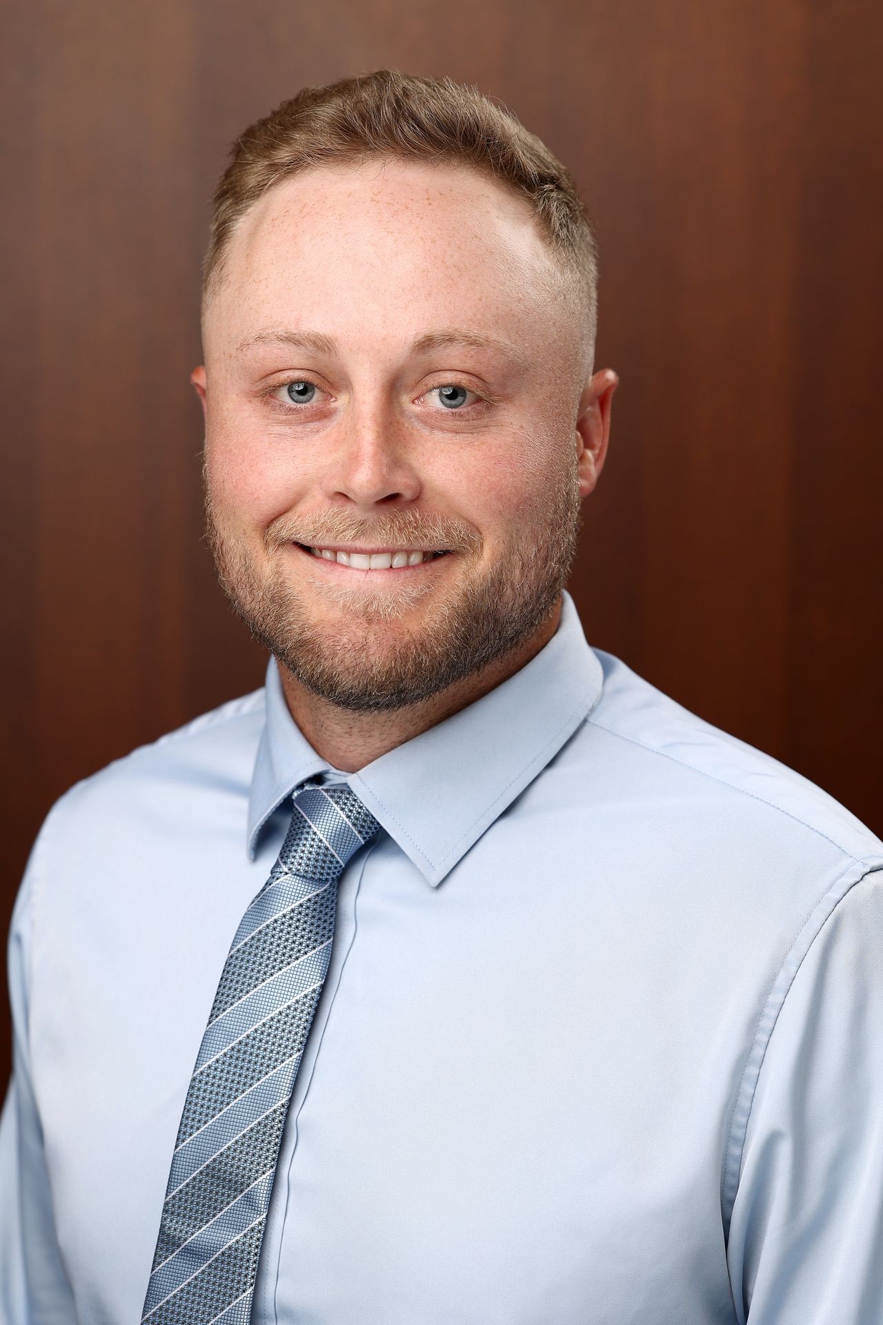 Man in a light blue dress shirt and patterned tie against a wooden background.