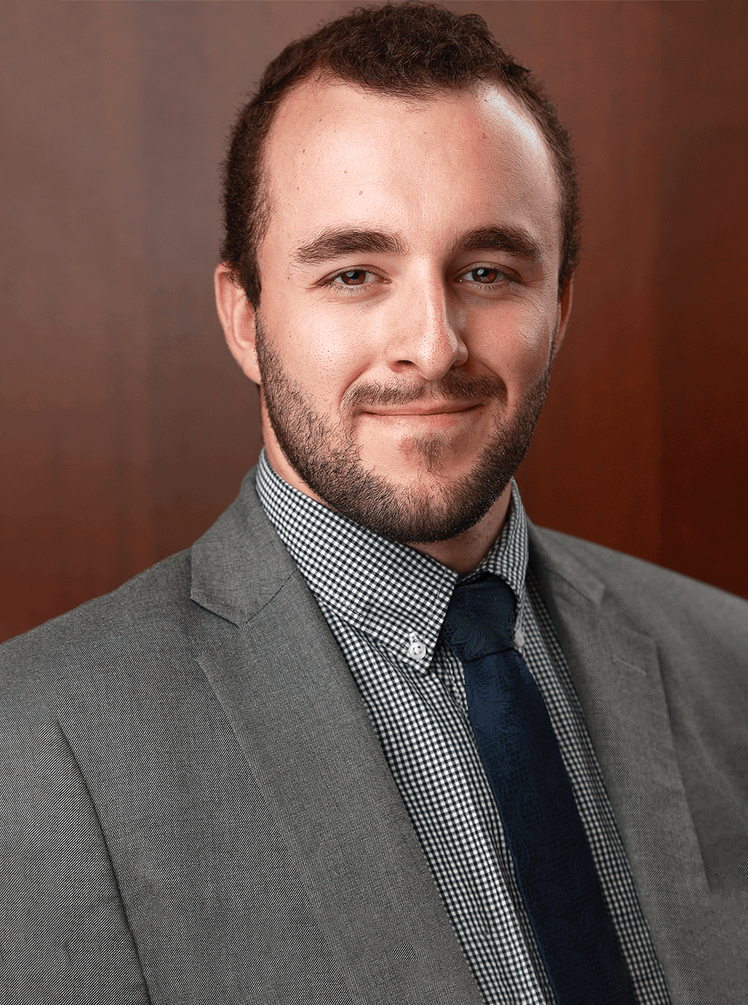 Man in a grey suit and checkered shirt with a navy tie smiling against a dark background.