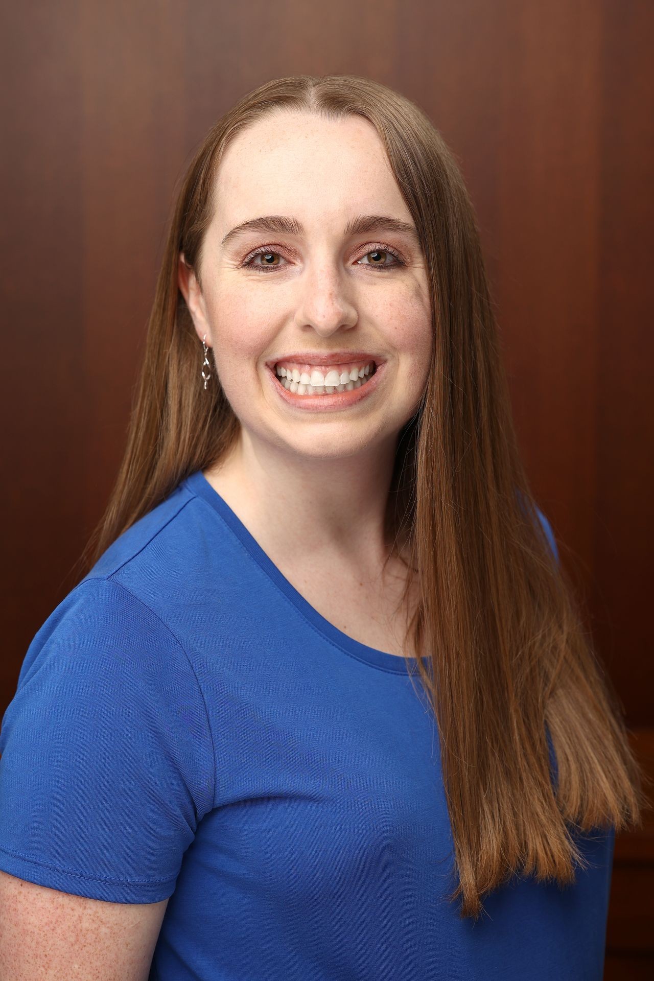 Woman with long straight hair wearing a blue shirt smiling broadly against a plain brown background.