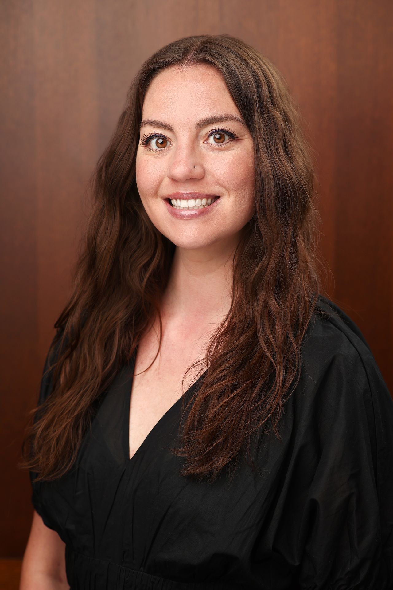 Person with long wavy hair wearing a black V-neck garment against a wooden backdrop.