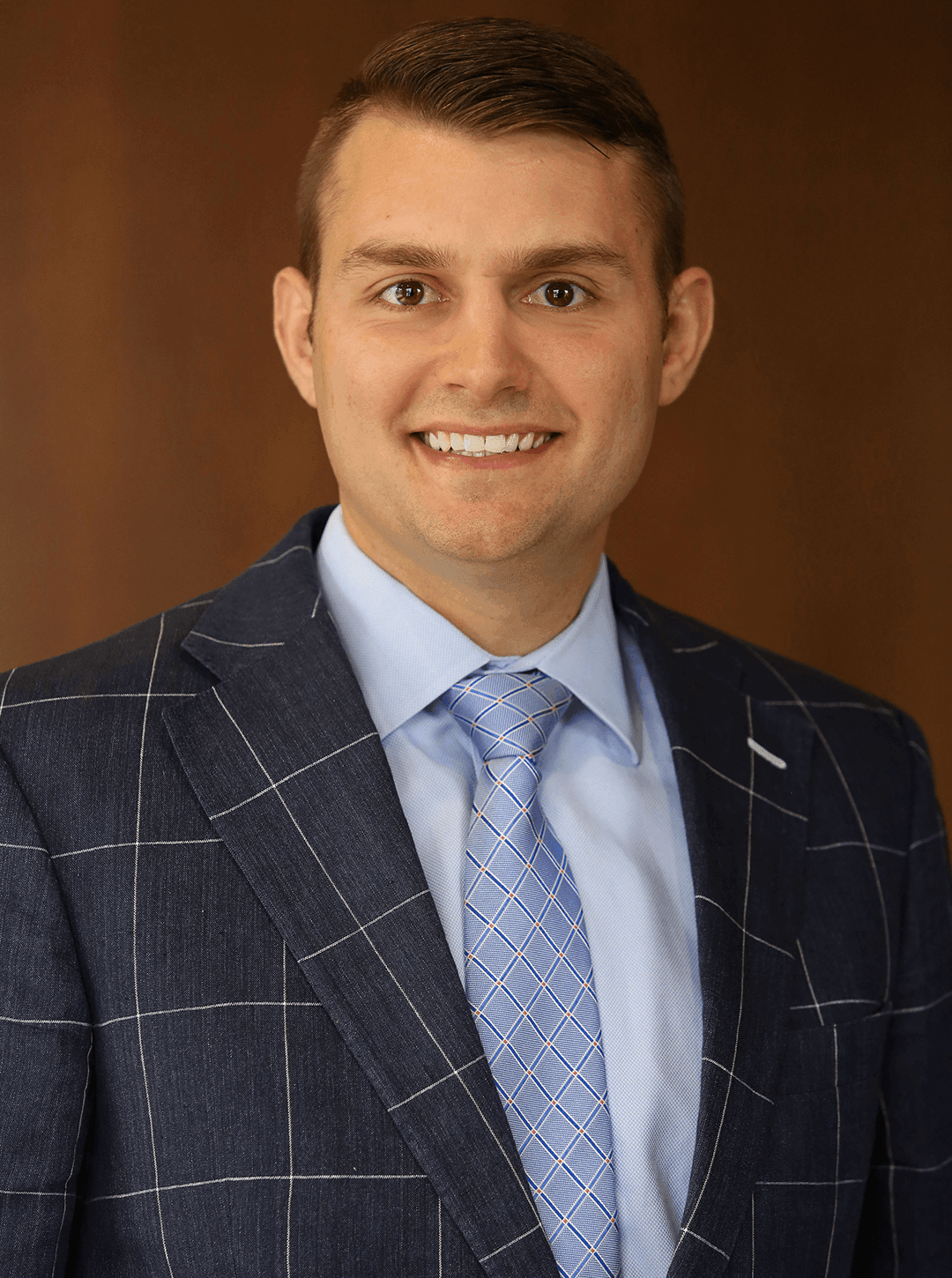 Man in a navy blue checked suit and light blue tie smiling against a brown background.