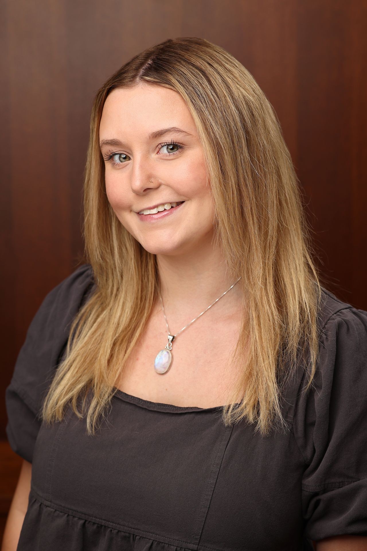 Person wearing a grey blouse and a necklace with a circular pendant, standing against a wooden background.